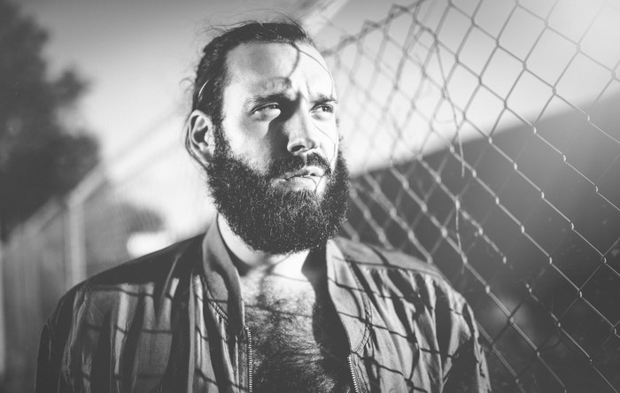 Man with beard leaning against a fence