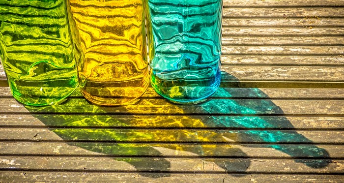 Three colored glass bottles casting shadows on wood planks