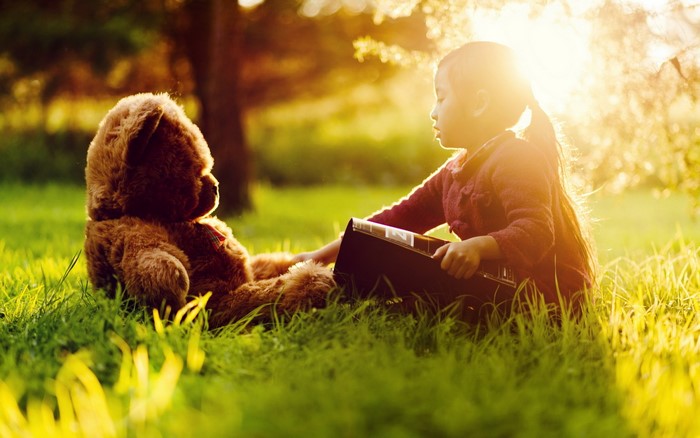 Girl reading a book to a teddy bear in the grass