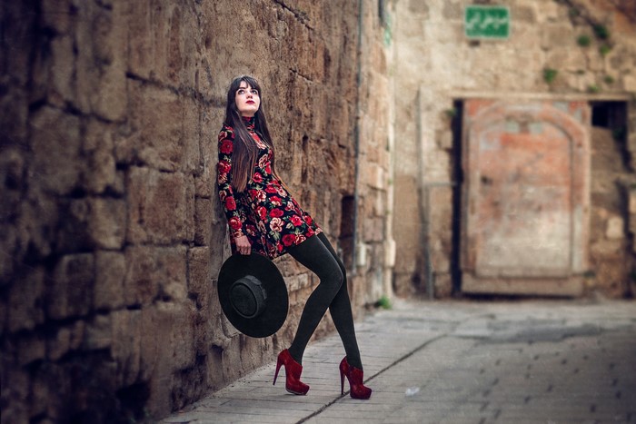 Woman in floral dress holding hat, leaning against stone wall