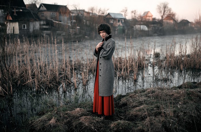 Woman in coat and dress by swamp with village