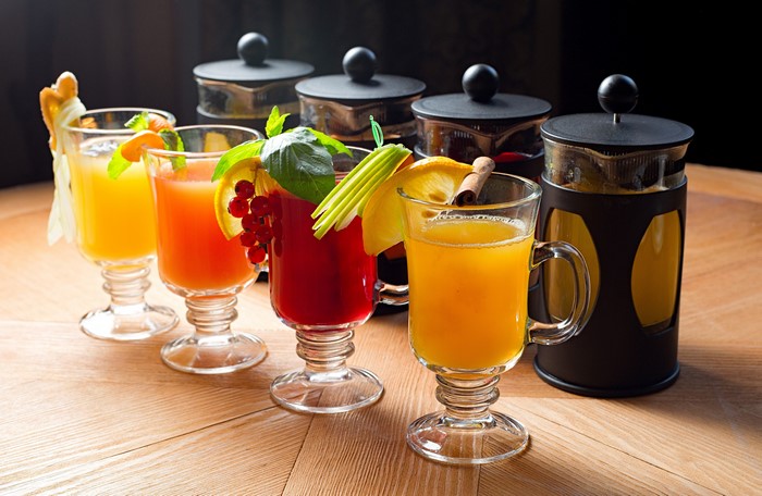 Four glasses of colorful drinks with garnishes and French presses