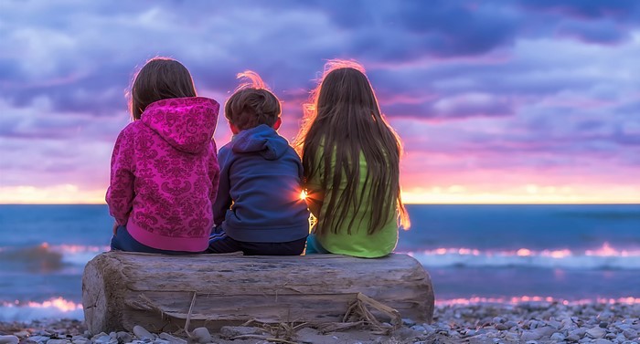 Three children sitting on a log at sunset