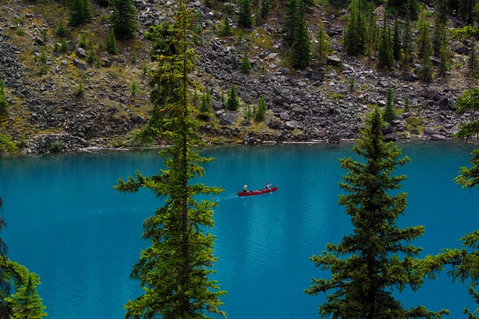 Two people in a red canoe on a bright blue lake with pine trees