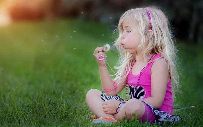 Blonde girl blowing dandelion seeds in grass