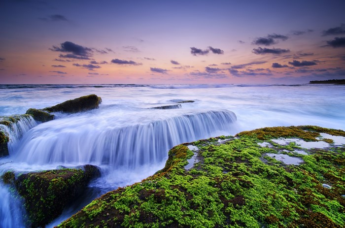 Waterfall over mossy rocks at Lima Beach, Bali at sunset