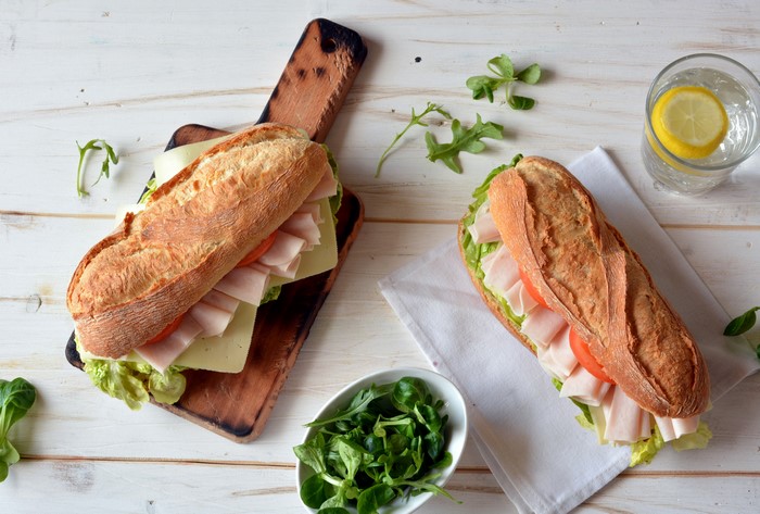 Two sandwiches with ham, cheese, lettuce, and tomato on a wooden board
