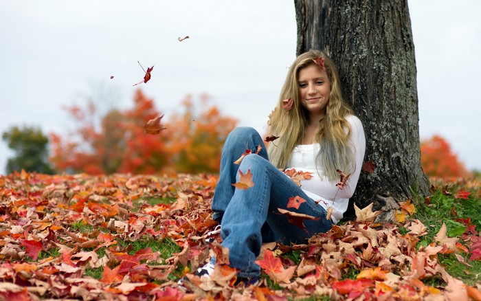 Blonde girl sitting in autumn leaves under a tree