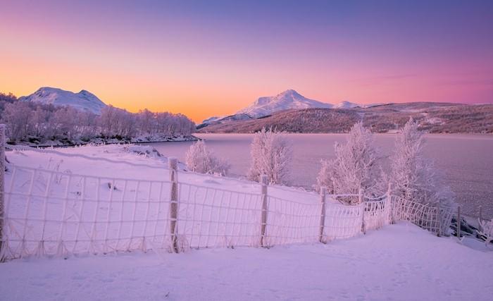 Winter sunrise over a frozen lake with snow-covered mountains and a fence