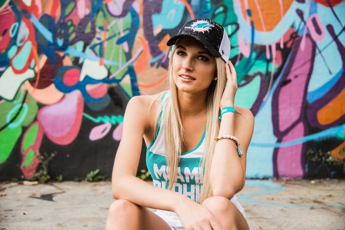 Blonde girl in Miami Dolphins hat sitting by graffiti