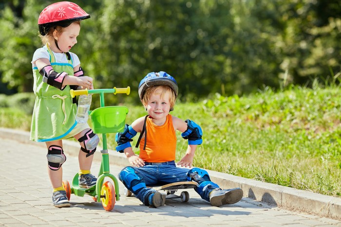 Two children with protective gear on a scooter and skateboard