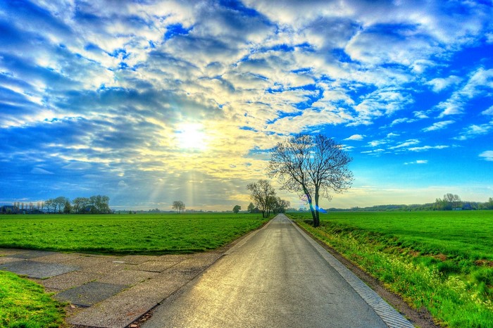 Road through green fields under a cloudy sky