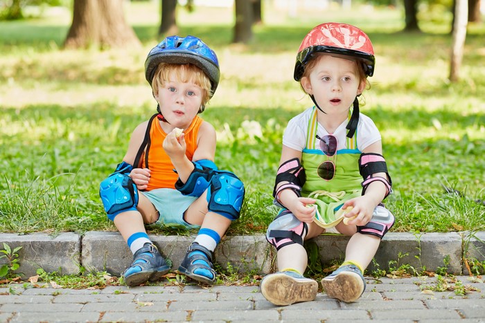 Two children wearing helmets and protective gear sitting on a curb