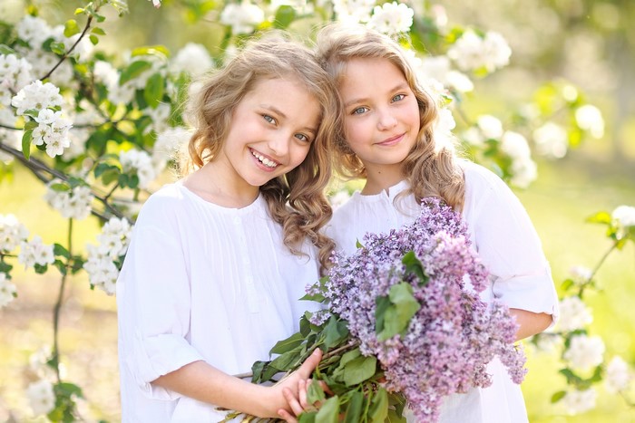 Two blonde girls holding lilacs in front of blooming tree
