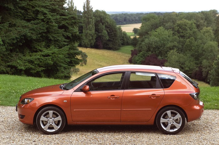 Orange Mazda 3 Hatchback parked on gravel with trees and fields