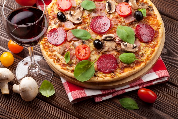 Pizza with Sausage, Mushrooms, Tomatoes, Olives, and Basil next to a glass of red wine