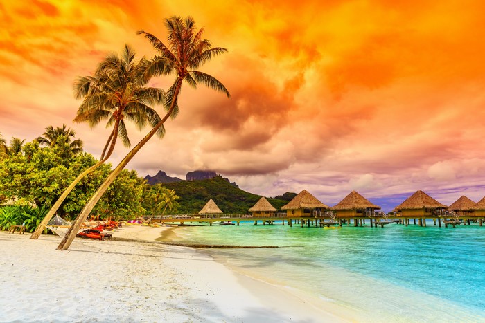 Tropical beach with overwater bungalows and palm trees at sunset