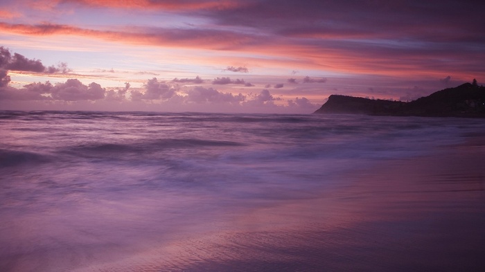 Ocean waves at sunset with colorful clouds and a distant hill