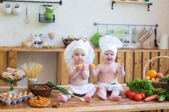 Two babies dressed as chefs in a kitchen with food