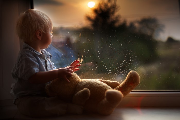 A toddler with a teddy bear looks out a window at sunset