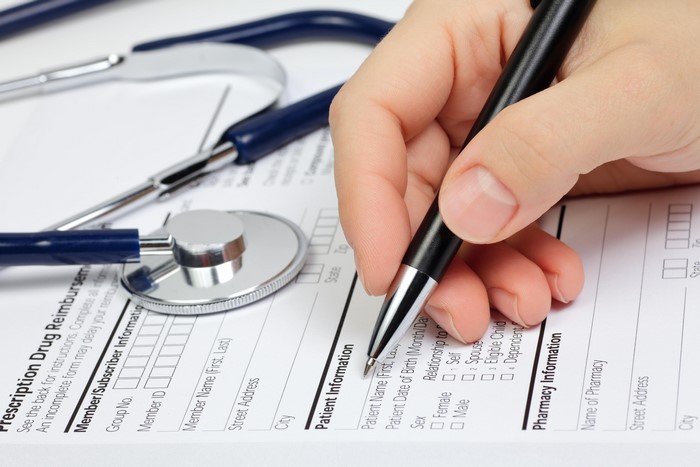 Hand filling out prescription drug reimbursement form with stethoscope