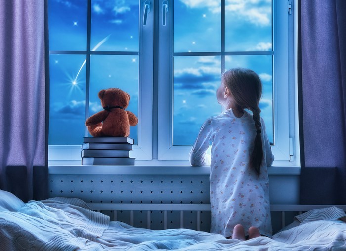 Little girl and teddy bear looking out window at night