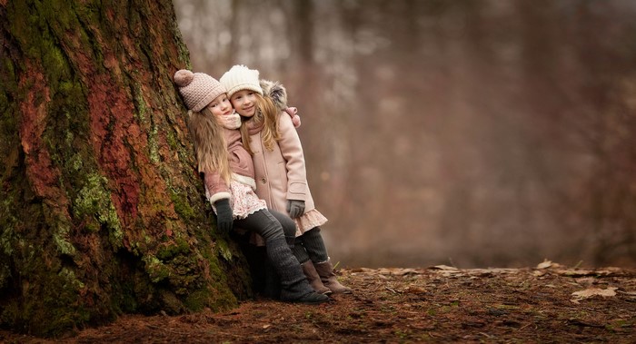 Two young girls in winter hats leaning against a tree trunk