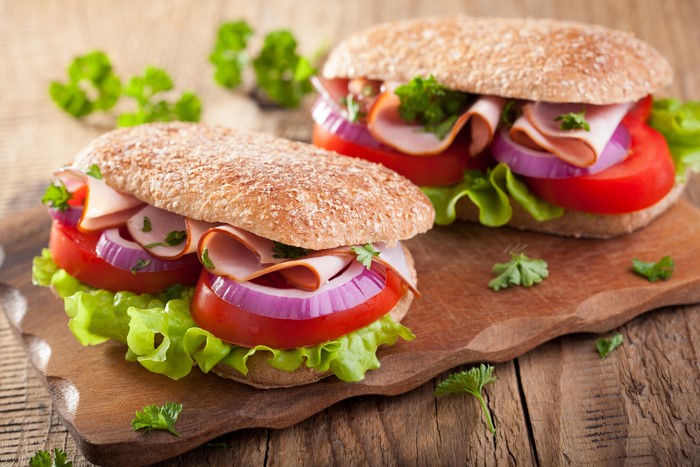 Two sandwiches with ham, lettuce, tomato, and onion on a cutting board