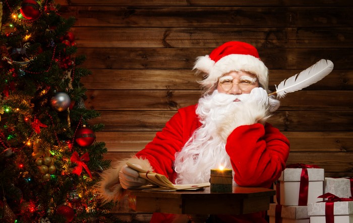 Santa Claus writing with a feather by a Christmas tree