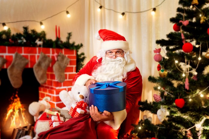 Santa Claus holding a gift box near a Christmas tree and fireplace