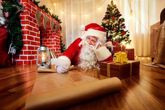 Santa Claus writing on a scroll by a fireplace and Christmas tree