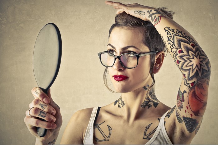 Woman with tattoos and glasses holding a mirror