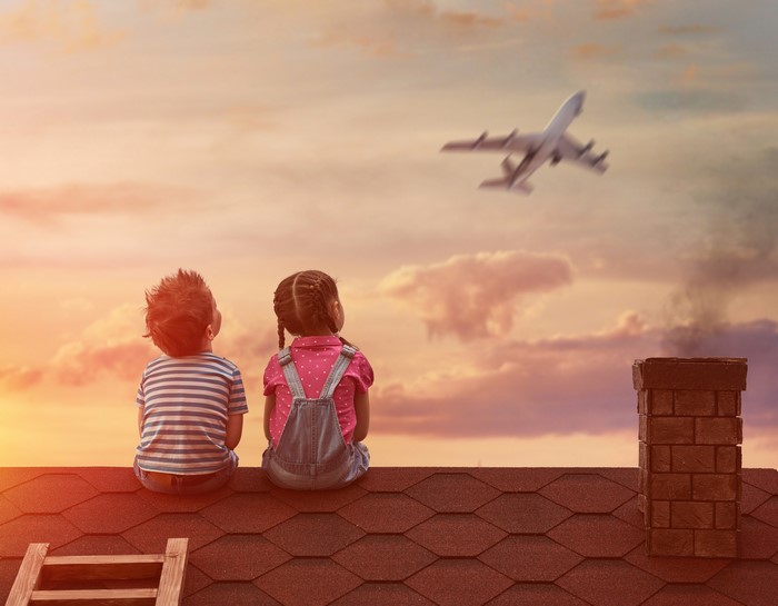 Two children sitting on a roof watching an airplane