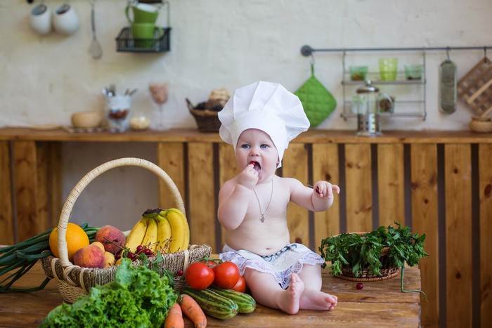 Baby in chef hat eating fruit in kitchen