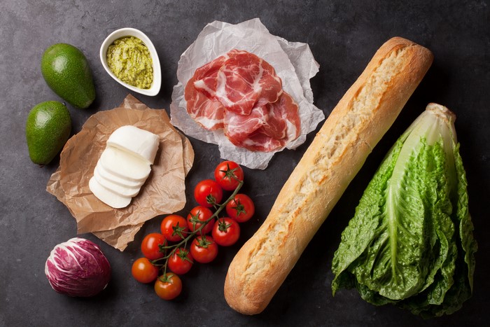 Still-life of sandwich ingredients: bread, ham, cheese, tomatoes, avocado, lettuce, radicchio, and pesto