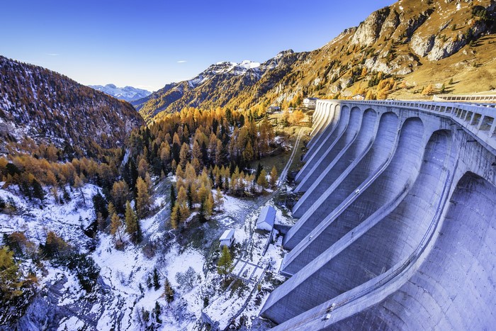 Curved concrete dam in a mountain valley with autumn trees and snow