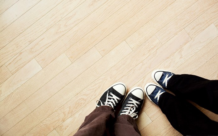 Two pairs of feet wearing sneakers on a wooden floor