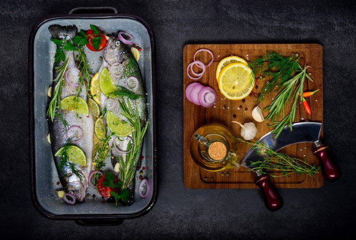 Two raw fish with lemon, lime, herbs, and spices in a baking dish next to a cutting board with ingredients