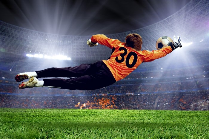 Goalkeeper in orange uniform jumping to catch a soccer ball in a stadium
