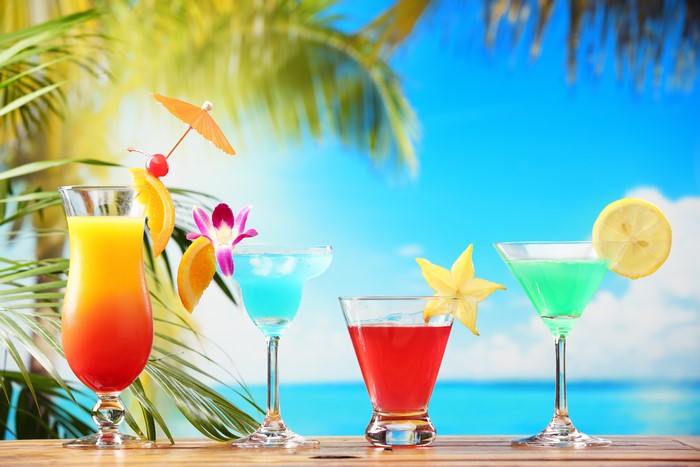 Four colorful cocktails on a wooden table with palm trees and ocean background