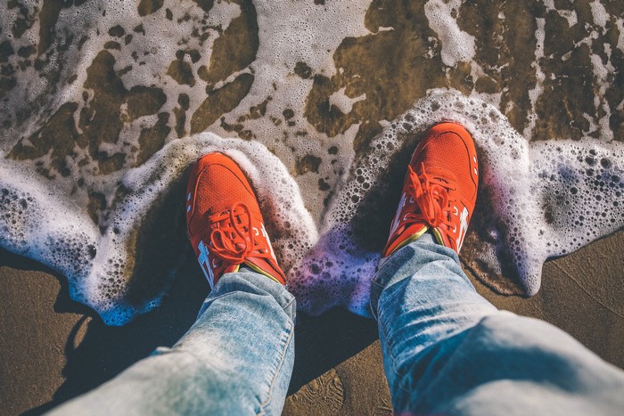 Person wearing orange athletic shoes and jeans on a beach with waves