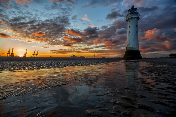 Lighthouse on a beach at sunset with industrial cranes