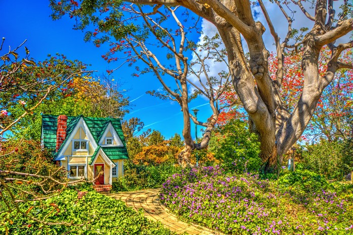 A charming miniature house in a vibrant garden with large trees.
