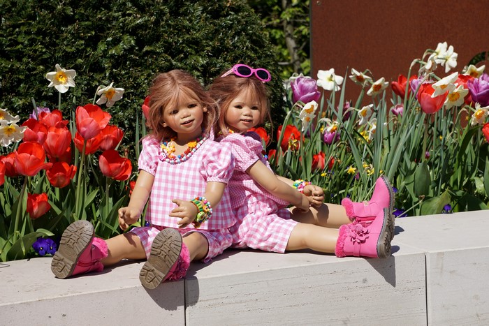 Two dolls wearing pink gingham outfits and boots sitting in front of flowers