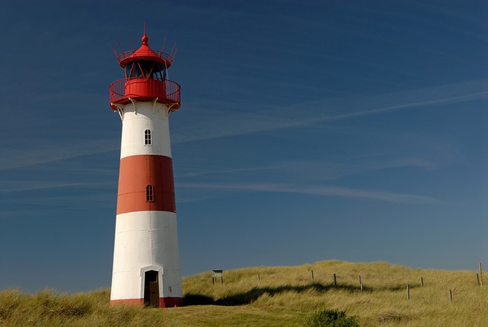 Red and white lighthouse on grassy dunes under blue sky