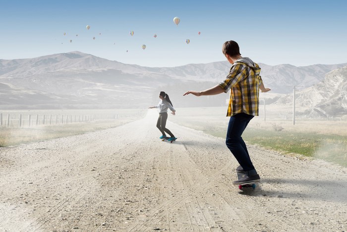 Two people skateboarding on a dirt road with hot air balloons