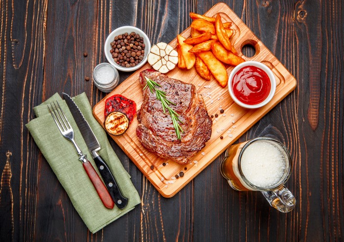 Steak with fries, ketchup, and beer on a wooden board