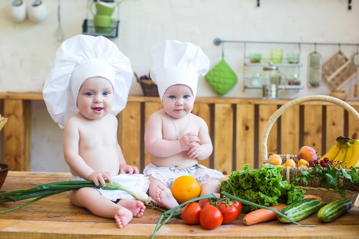 Two babies dressed as chefs in a kitchen with vegetables