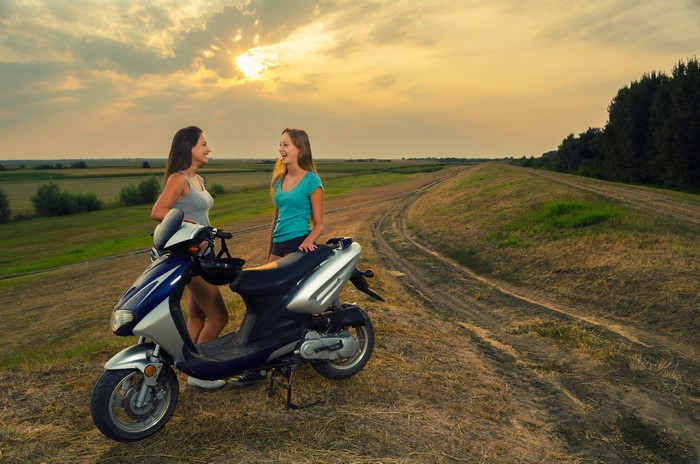 Two young women and a scooter on a dirt road at sunset