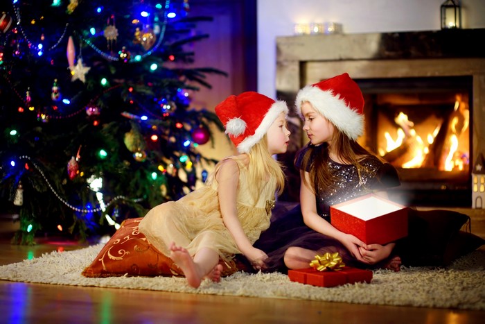 Two girls in Santa hats opening a Christmas gift by the fireplace and tree